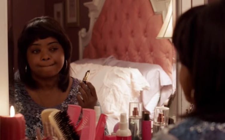 A woman sits at a vanity table applying lipstick.