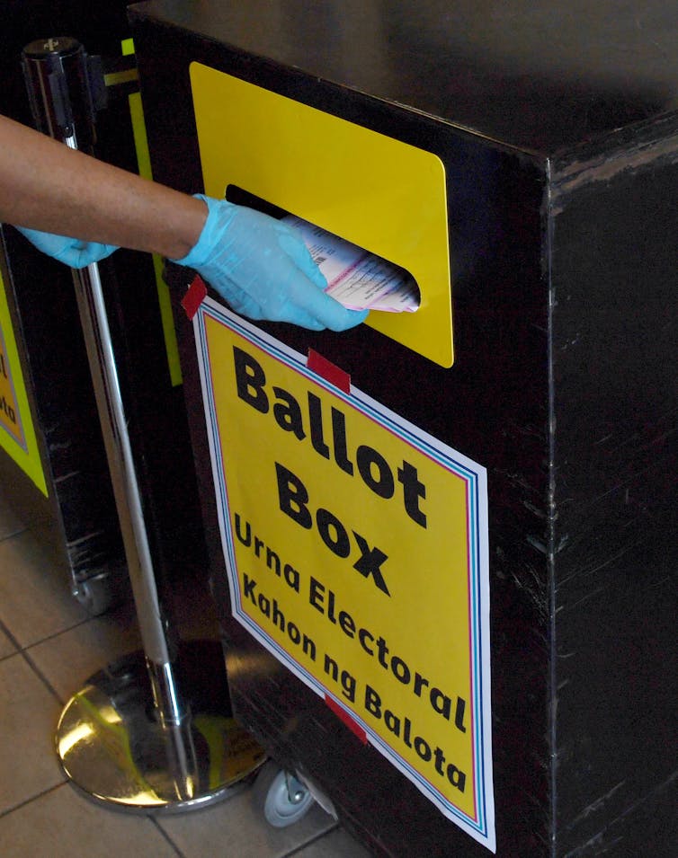 A gloved hand inserts papers into the slot of a black and yellow box labeled Ballot Box