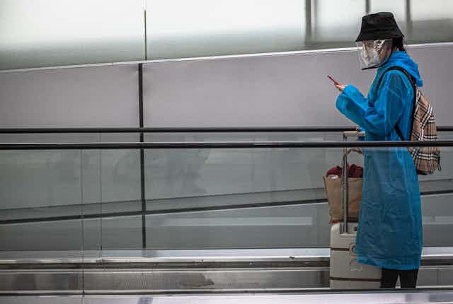 Una mujer con maletas, sombrero, mascarilla y pantalla facial mira el móvil en una cinta transportadora de nodo de transportes.