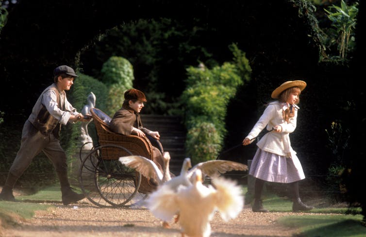 A girl walks ahead of a boy pushing another boy in a wheelchair. Geese flap in their wings in the forefront.