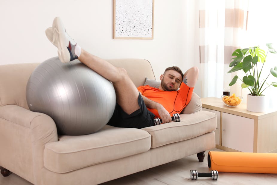 Young man is laying on a couch wearing exercise clothes, resting his feet on an exercise ball.