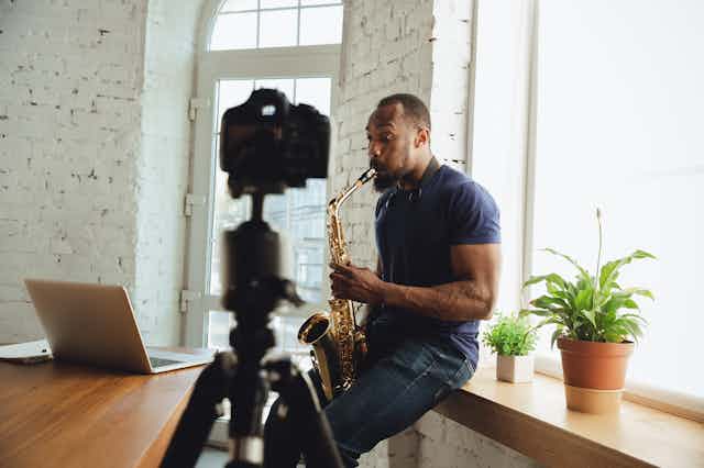 Un saxofonista toca el instrumento ante una cámara y un ordenador.