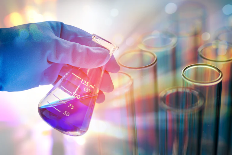 Light shines from behind a gloved hand holding a beaker containing a blue liquid with test tubes in the background