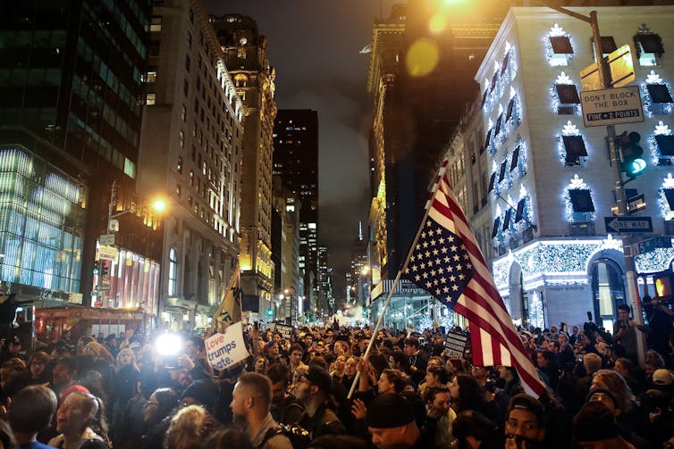 Protesters in NYC at night, holding anti-Trump signs and waving American flags