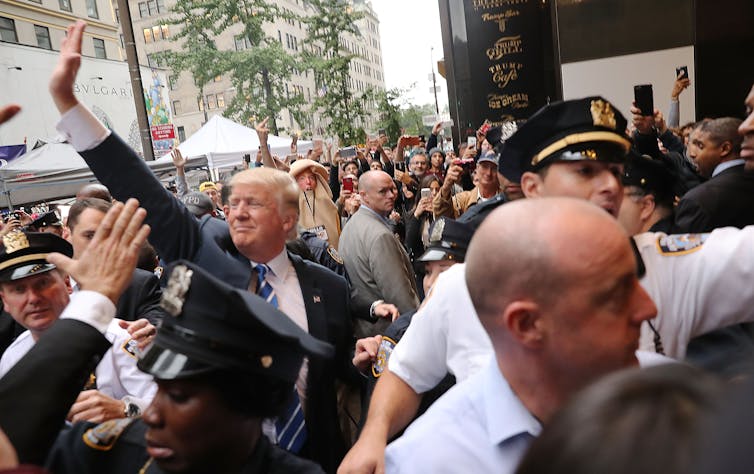 Trump, surrounded by police, waves to supporters in a crowd with protesters in the background