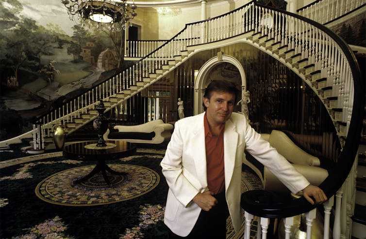 Trump in a high-ceilinged foyer with dramatic wallpaper, rug and staircase