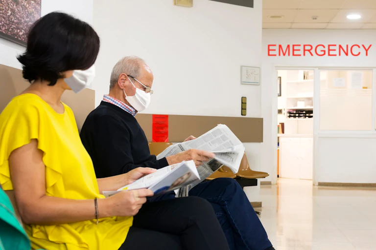 A younger woman and an older man wearing facemasks sit in a waiting room at a hospital.