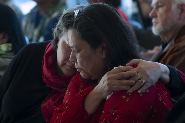 Two seated women embrace with their hands resting on one woman's shoulder.