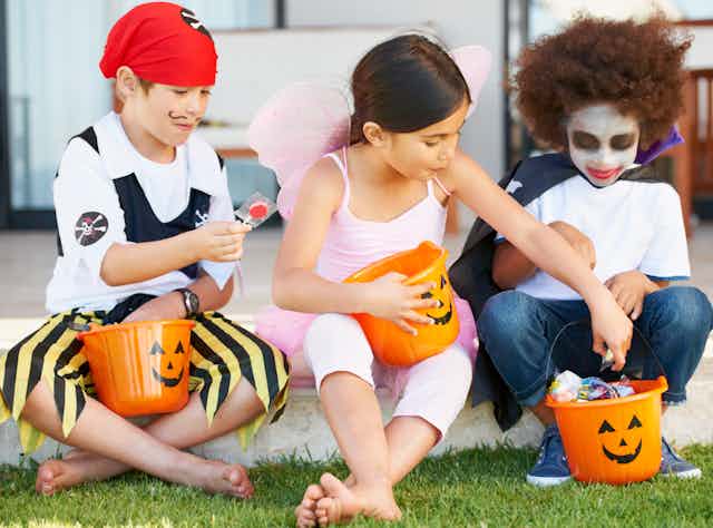 Three children dressed in Halloween costumes holding candy buckets while sitting down.
