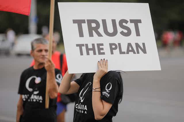 A women in a black tshirt holding a sign reading TRUST THE PLAN