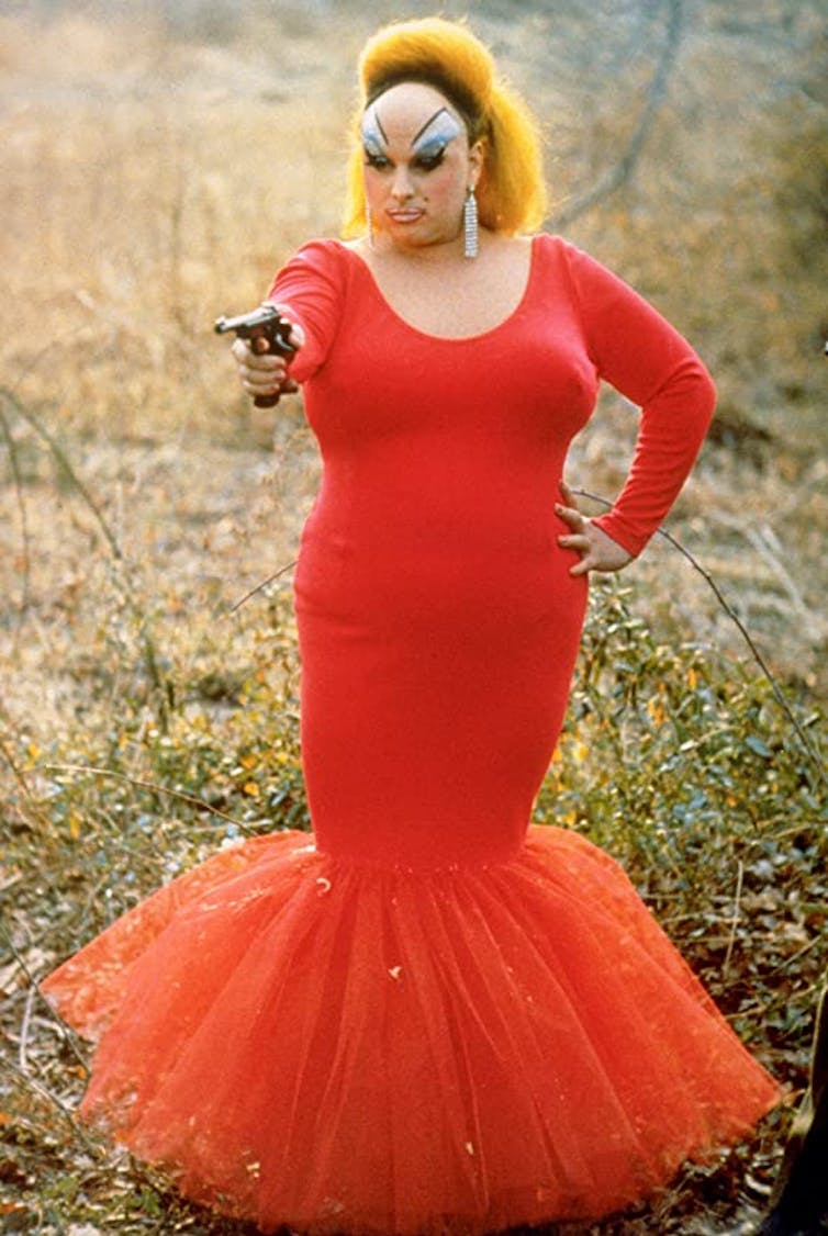 Devine wearing a red dress, holding a gun