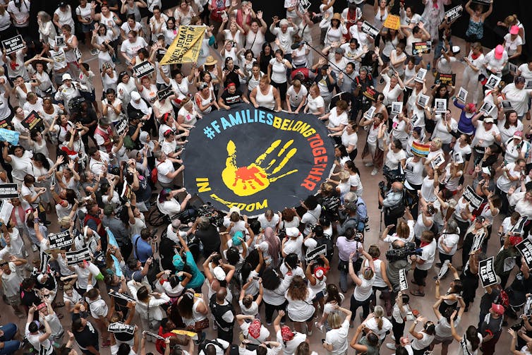 A crowd of women in white hold up a giant banner reading #FamiliesBelongTogether