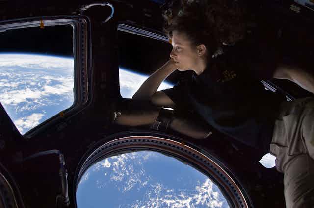 A woman floats in front of windows showing a view of Earth far below.