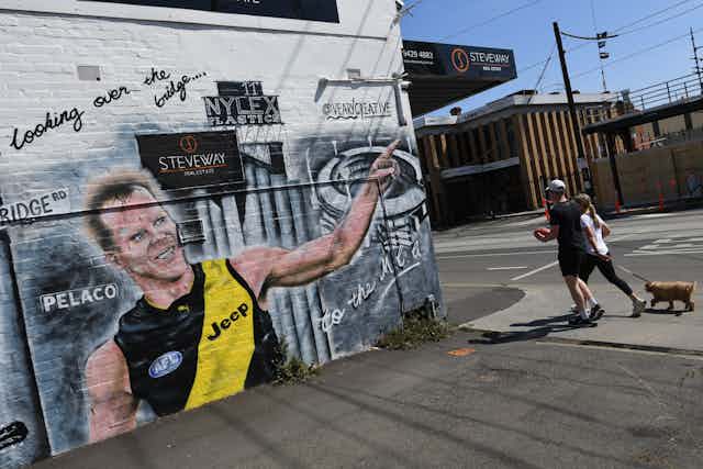 Two people walk past a mural in Melbourne depicting a Richmond footballer celebrating.