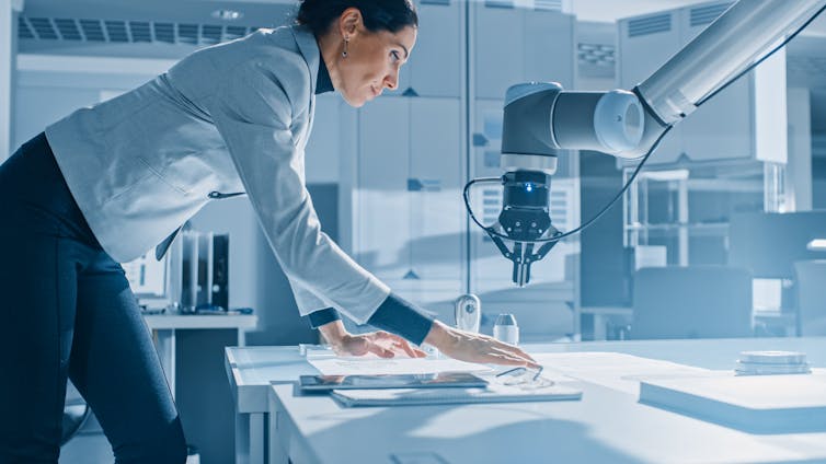 Female robotics engineer leans on the table as she works with blueprints, documents and tablet computer to program robot arm movements.