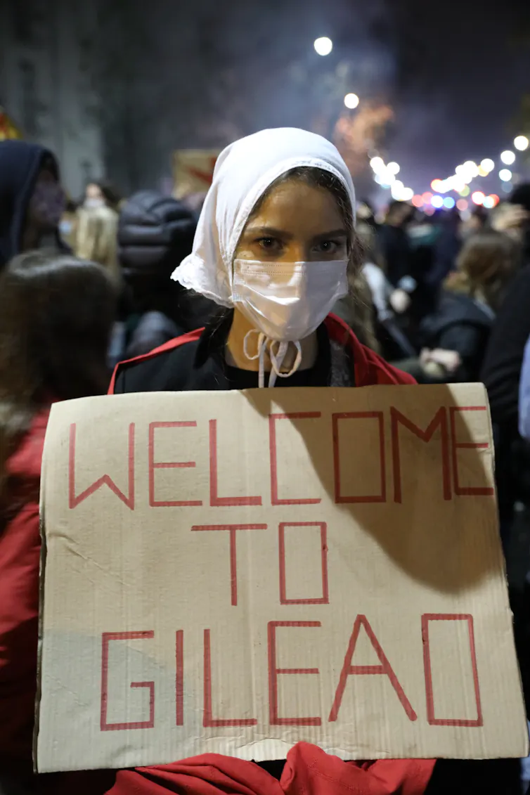 Women in Handmaid's Tale costume holds sign reading 'Welcome to Gilead'.