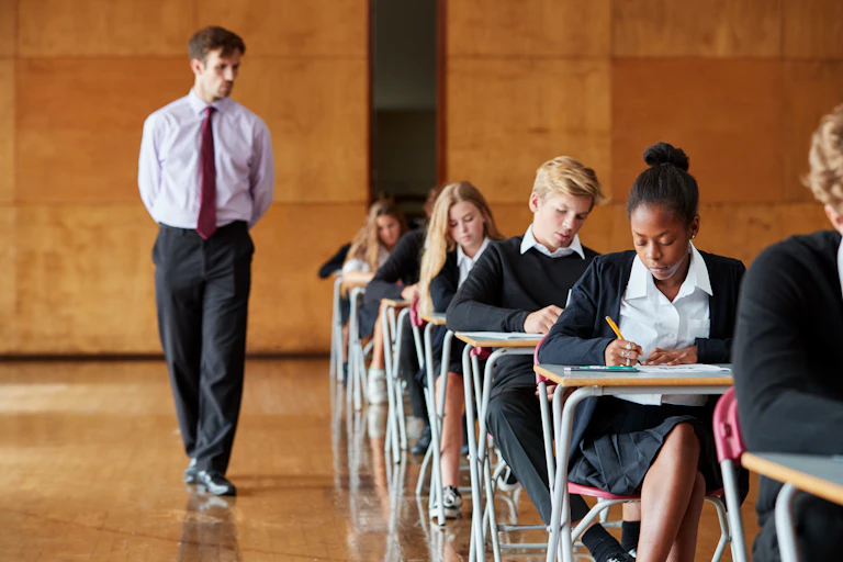 Teenage students writing an exam with a teacher invigilating.