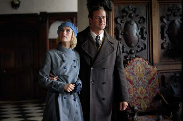 Woman and man in 1940s costumes stand in the entrance hall of a stately home.