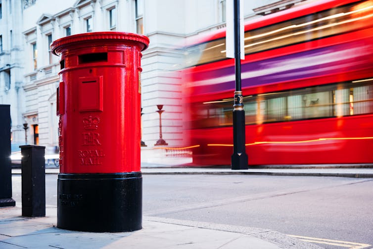 Red post box on street with red London bus behind.
