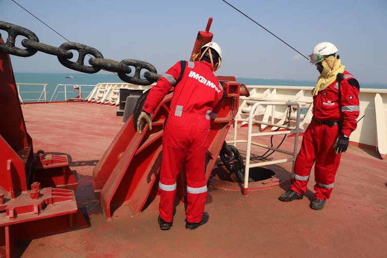 Two people in high visibility suits work on a ship deck.