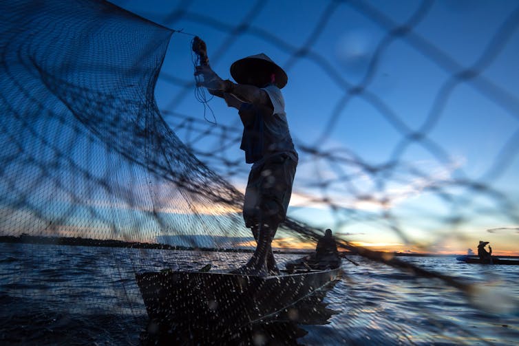 A fisher on a wooden boat casts a net into tropical water at dusk.