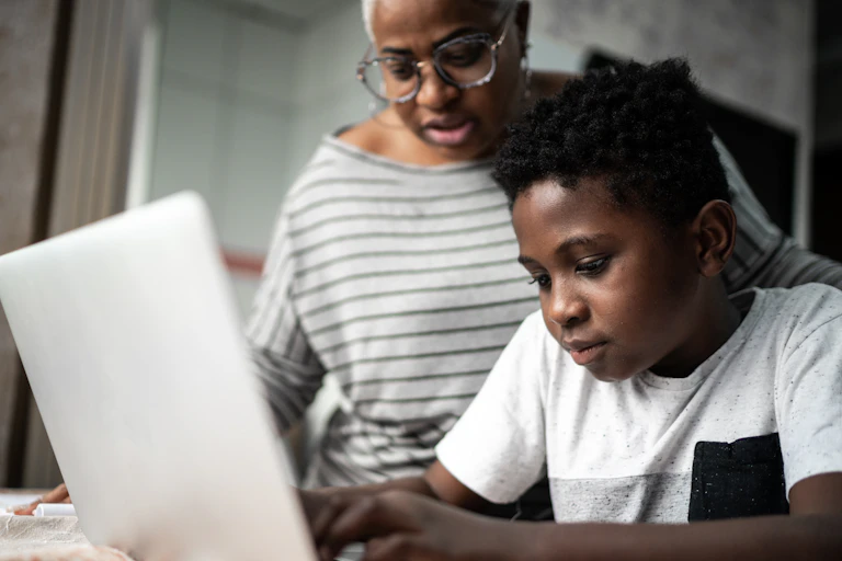 A mother watches as her son works on a laptop at a table in their home.