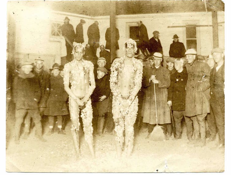 Two African American men stand together doused in tar and feathers among a crowd of white students .