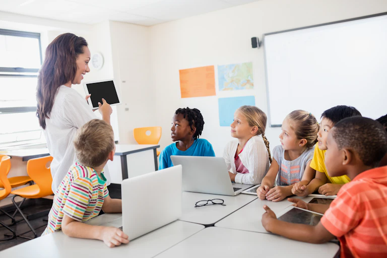 A primary school teacher pointing to a ipad with students looking on.