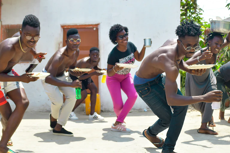 Seven young men and women dance in a backyard holding plates of food in a line dance formation, many of them smiling and having fun.