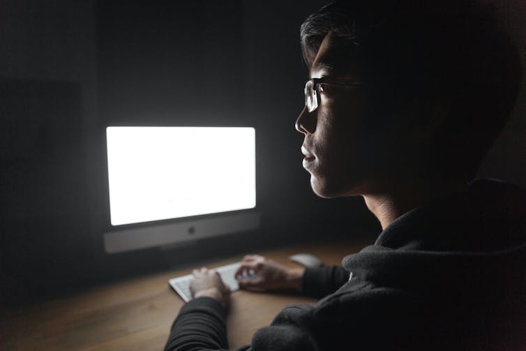 Young man with glasses sitting in a dark room in front of his computer screen.