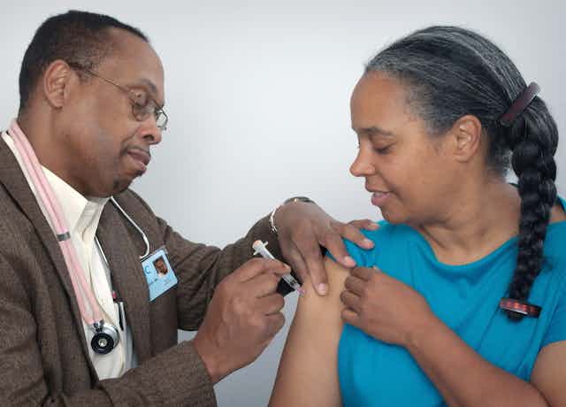 A man wearing a stethoscope gives a woman a shot in her upper arm.