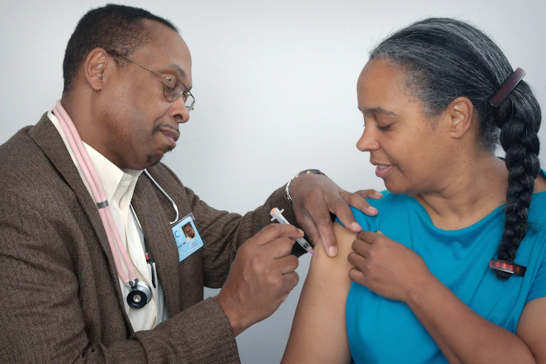 A man wearing a stethoscope gives a woman a shot in her upper arm.