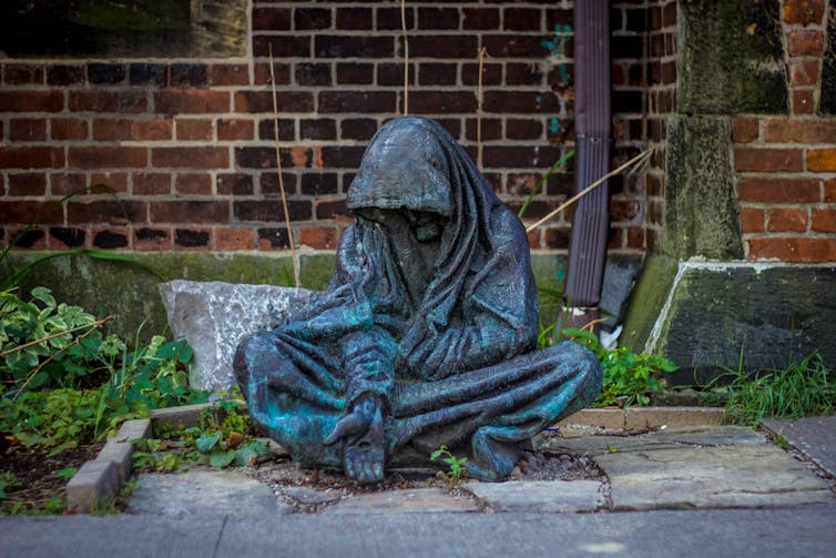 a bronze sculpture depicts a person wrapped in a blanket with an hand stretch out palm turned up as if asking for food or money.