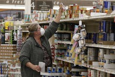 An older woman in a mask reaching to grab a can of food at a grocery store.