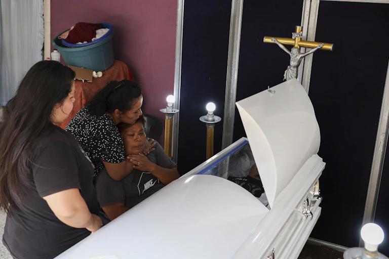 A weeping mother sits beside a white casket as two women stand near one, one of them embracing her.