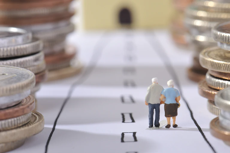 Una pareja de muñecos de ancianos camina de espaldas entre monedas apiladas en lo márgenes de un camino dibujado en el suelo.
