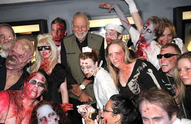 Zombies surround man in a green vest and a white beard.