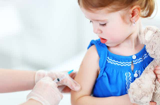Child clutching teddy bear watching as health professional injects upper arm