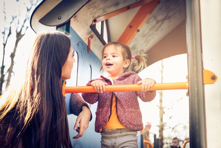 A young girl on equipment in the playground with her mother.