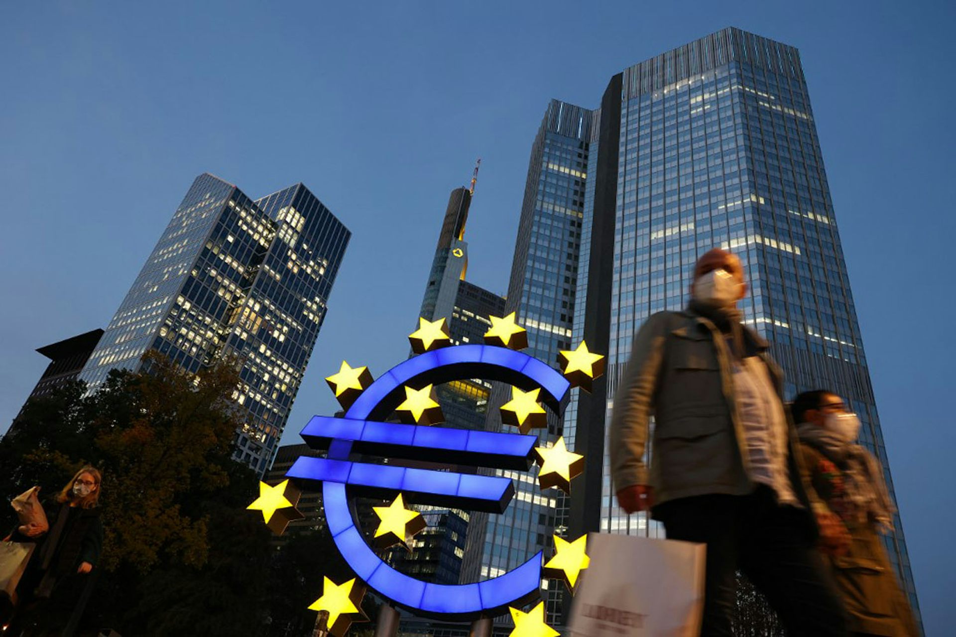 People wearing face masks walk in front of a euro sign in the center of Frankfurt am Main, Germany