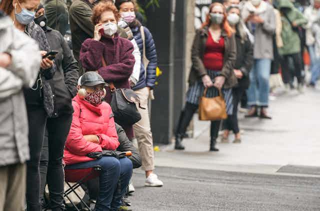 People wearing masks and one sitting in a folding chair wait in a long lineup along a city street.