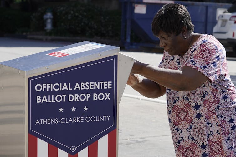 A woman in a flowered shirt puts her ballot into a ballot box outdoors.