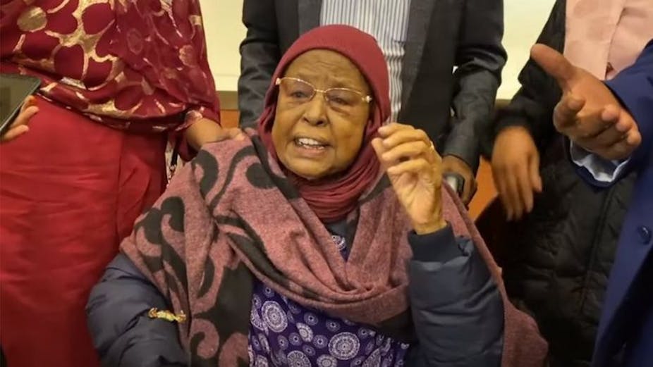 An older woman in a headscarf and wearing glasses talks with her hand raised.