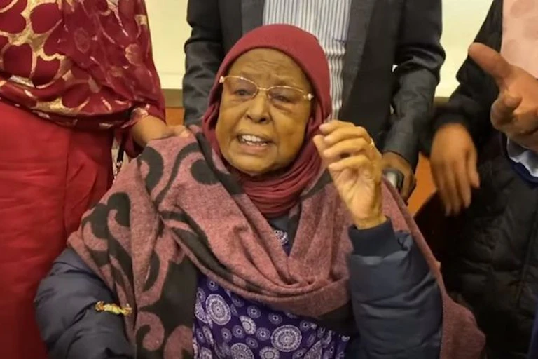 An older woman in a headscarf and wearing glasses talks with her hand raised.