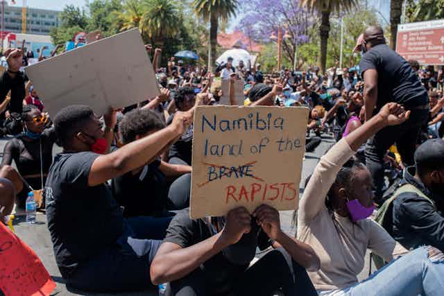 Protesters, seated on a city road, wearing face masks and holding up fists, in the foreground a protester holds a placard that reads, Namibia land of the brave - the brave crossed out and replaced with the word rapists.