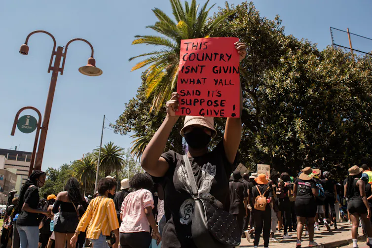 A woman dressed in black and wearing a face mask holds up a red poster that reads, 'This country isn't given what y'all said it's suppose to give.'