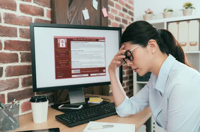 A woman with eyes closed and hand on forehead sits in front of a computer screen showing a ransomware demand