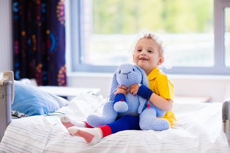 Young happy child sits on hospital bed holding a blue bunny toy.