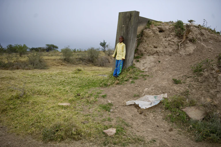 A boy wearing a long-sleeved yellow shirt and green long pants leans his back against an abandoned concrete structure on a farm in South Africa.