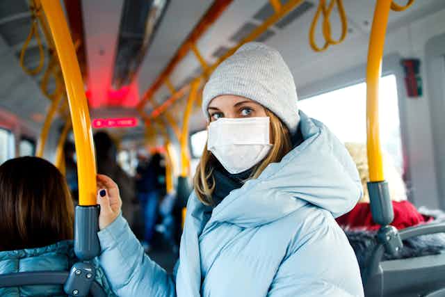 Woman wearing face mask, coat and hat, standing inside a bus